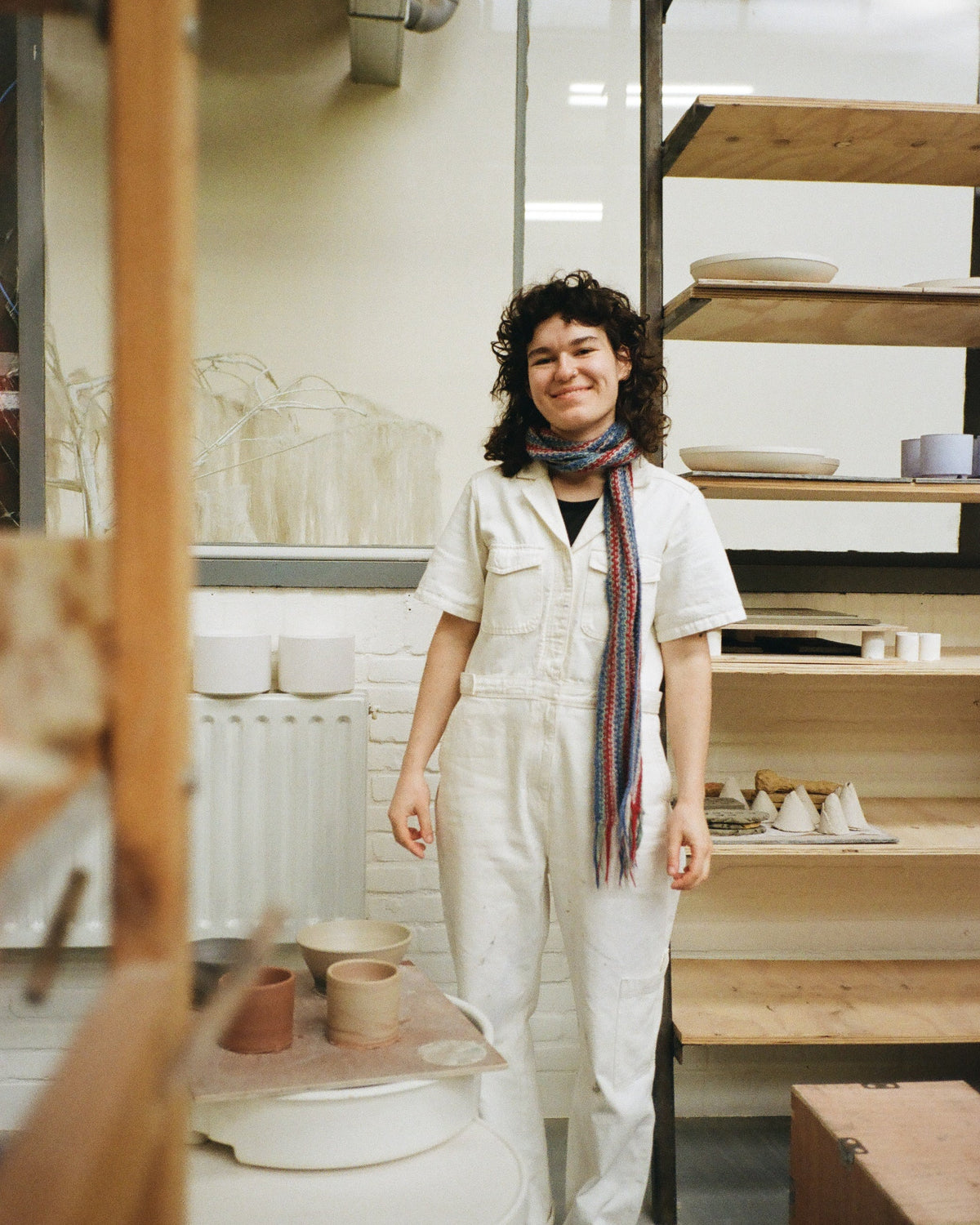 A woman with curly dark hair stands in a pottery studio wearing a white short-sleeve jumpsuit and a striped scarf. Behind her are shelves with ceramic plates and pottery on a wheel.